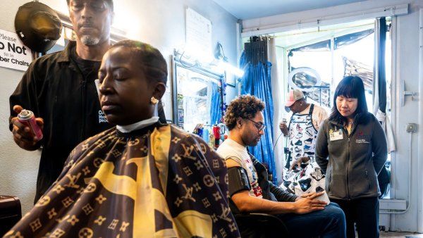 Barber cuts hair of woman in chair while Zhou interviews other man in chair.