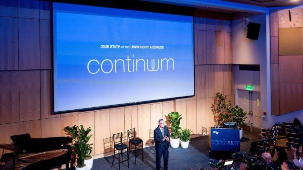 Chancellor Hawgood stands in front of audience and camera with the words "continuum" on the projector screen.