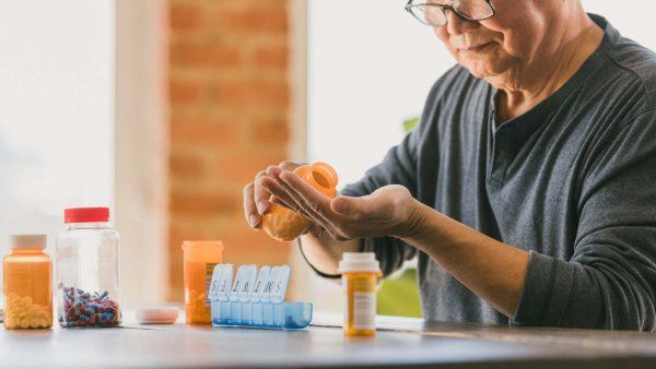 Elderly person with glasses pouring prescription pills from an orange bottle into their hand next to a weekly pill organizer.