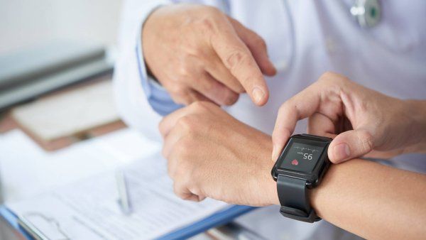 A doctor points to a person's wrist wearing a smartwatch displaying a heart rate of 95 bpm, with paperwork in the background.