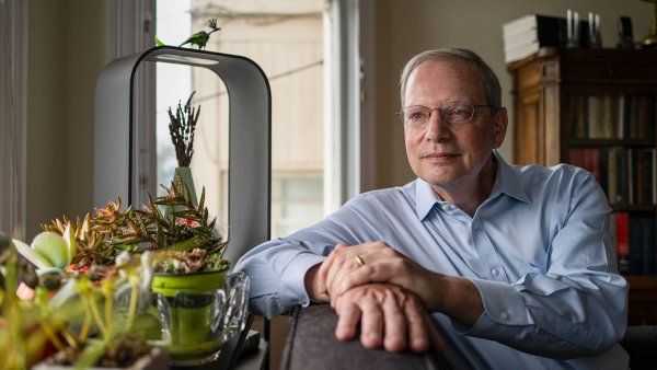 Robert Wachter sits on his couch in his home, resting is arm on the back with succulent pants in the foreground by the window.
