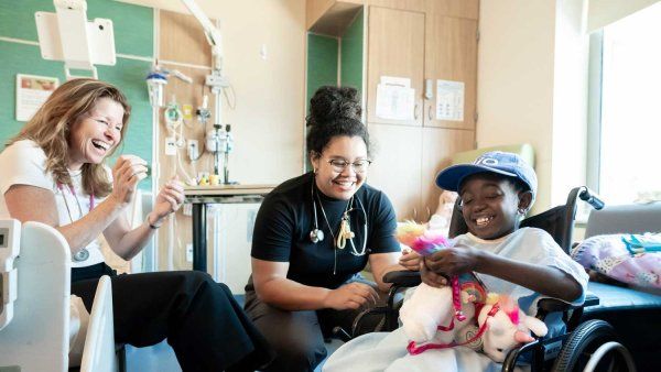 Pediatricians Michele Long and Kathleen Wallace sit with a young patient from Tanzania who sits in a wheelchair.
