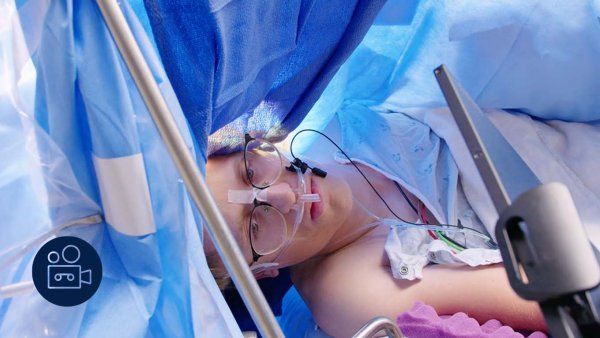 A young man in glasses has his eyes open during surgery as he looks at a screen. Blue surgery drapes are all around him.