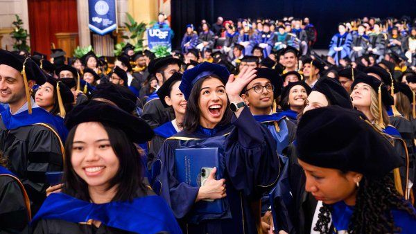 A large group of graduates in blue and black academic regalia at a U C S F commencement ceremony, with one graduate smiling and waving while holding a diploma folder.
