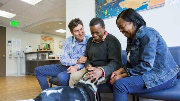 Doctor Ed Machtinger meets with two Black female patients, who smile as they interact with a therapy dog.