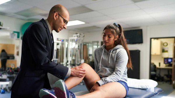 Orthopedic surgeon Nirav Panya examines a surgical scar on a patient's knee inside a physical therapy room.