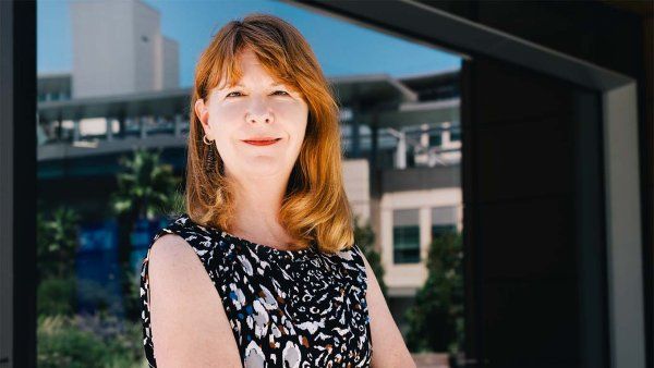 Joanne Spetz stands with her arms folded in front of a large window reflecting UCSF's Mission Bay campus.