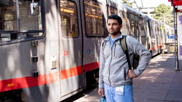 A medical student stands in front of a San Francisco MUNI rail train.
