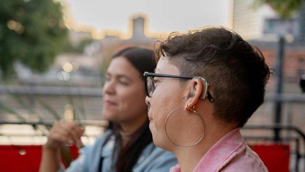 A woman wearing a hearing aid sits outdoors with a companion.
