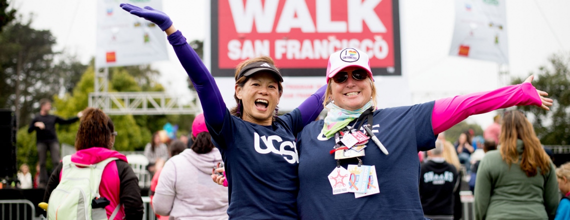 2 women wearing UCSF t-shirts and pink tutus pose in front of the AIDS Walk sign