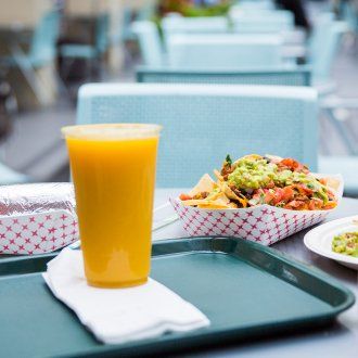 A large orange juice and a container of nachos piled with fresh guacamole and pico de gallo on a tray