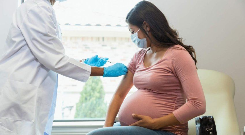 A doctor wearing a white coat and blue latex gloves injects a vaccine into a pregnant woman's arm.