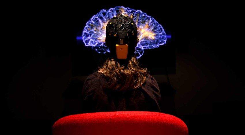 A person seen from behind sitting in a red chair and wearing a black EEG head-cap with numerous yellow sensors and wires. They are looking at a large screen in a dark room that displays a glowing, translucent blue and orange 3D visualization of a human brain.
