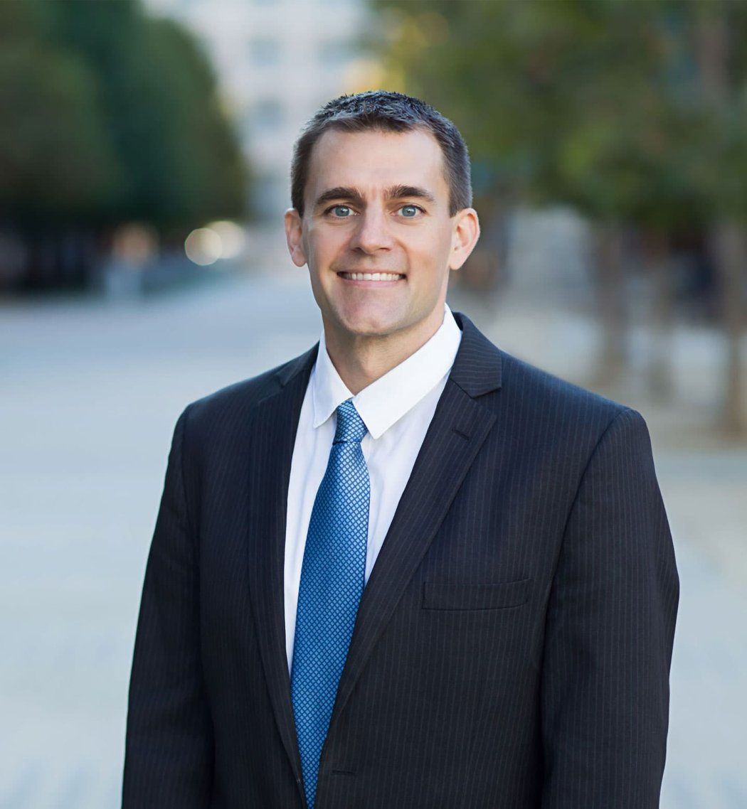 Jon Kleen wears a black suit and blue tie as he poses for a portrait. Trees are in the background.