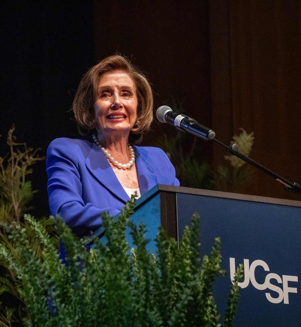 Nancy Pelosi stands onstage at a podium that has a sign that reads "UCSF."