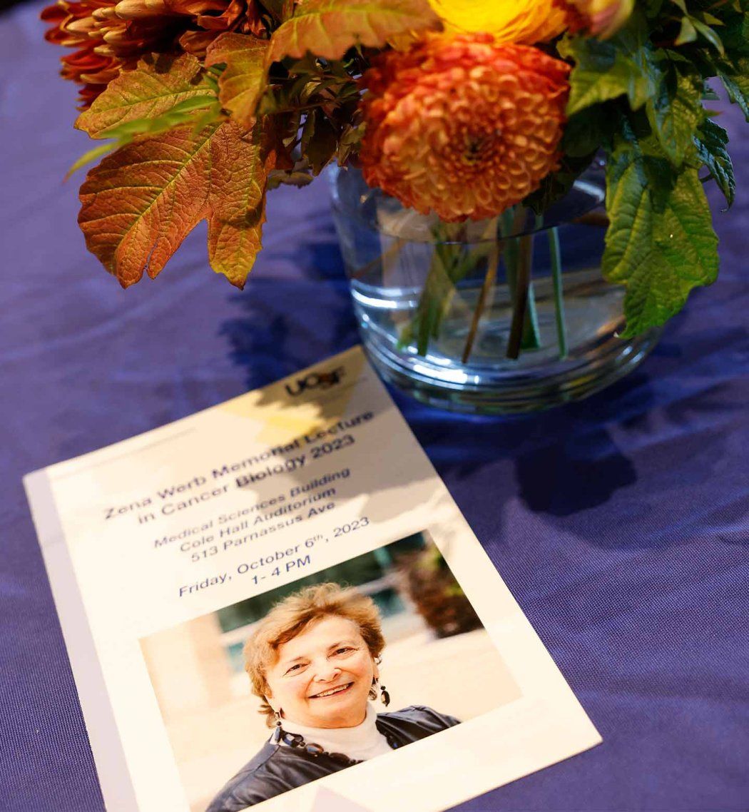 A leaflet that reads "Zena Werb Memroial Lecture in Cancer Biology 2023" and also includes the date and location of the 2023 event. The leaflet is rested on a vase of colorful flowers.