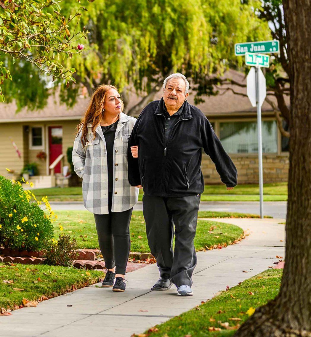 A Hispanic woman walks arm-in-arm with her elderly father along a grassy sidewalk in Salinas, California.