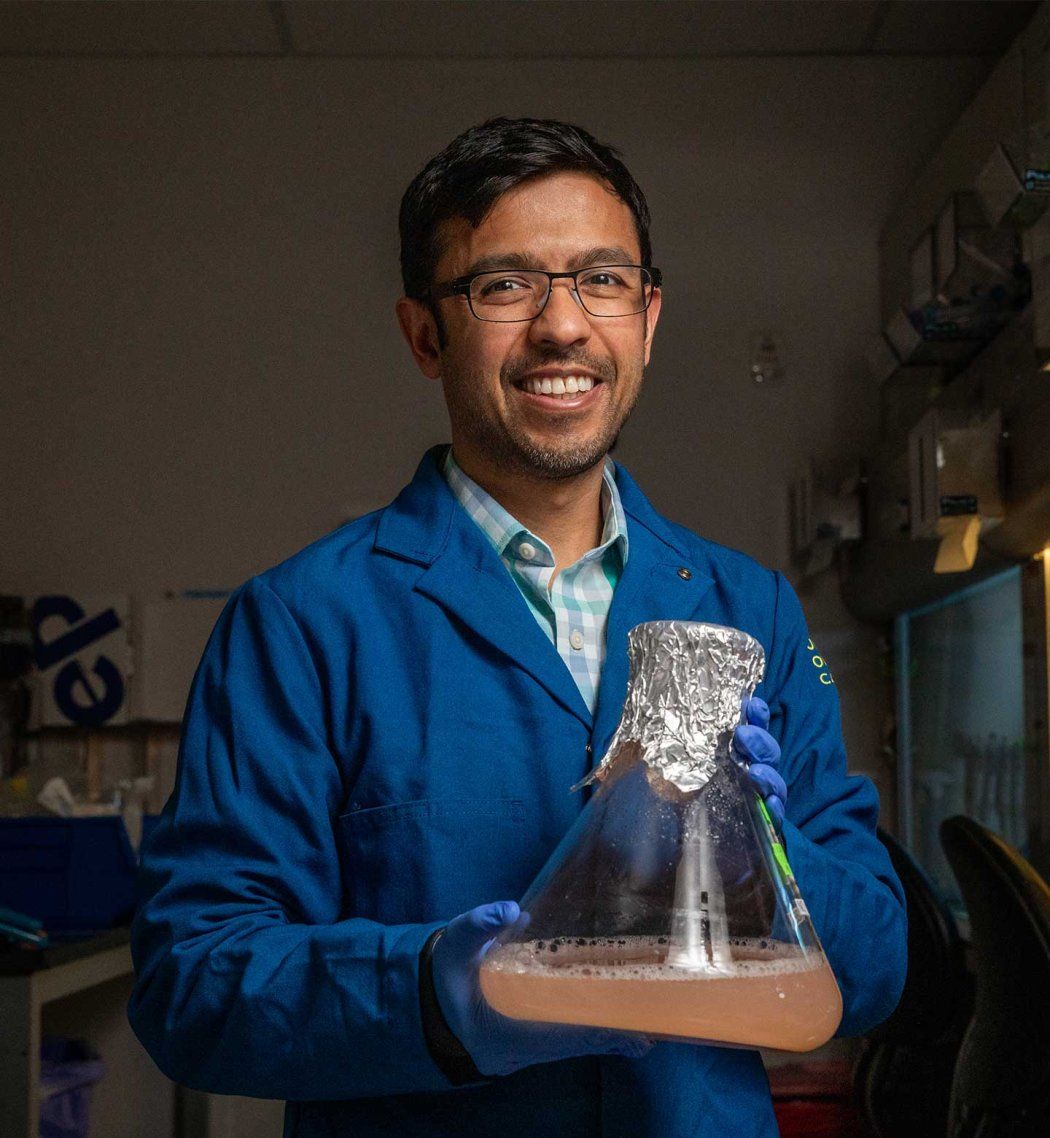 Aashish Manglik wears a blue lab coat and holds a large chemical flask with a pink liquid.