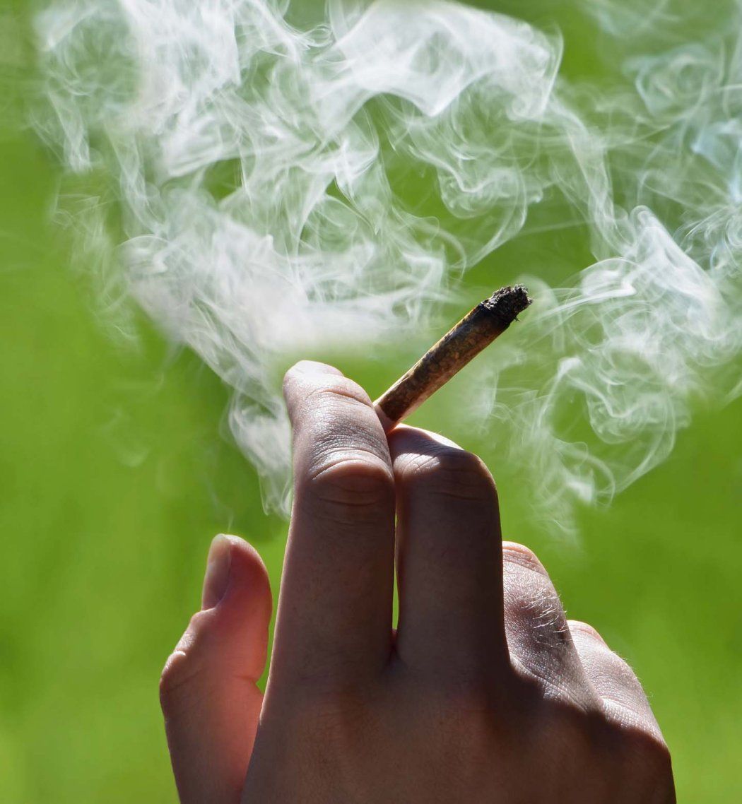A person holds a smoking joint between their fingertips against a green background.