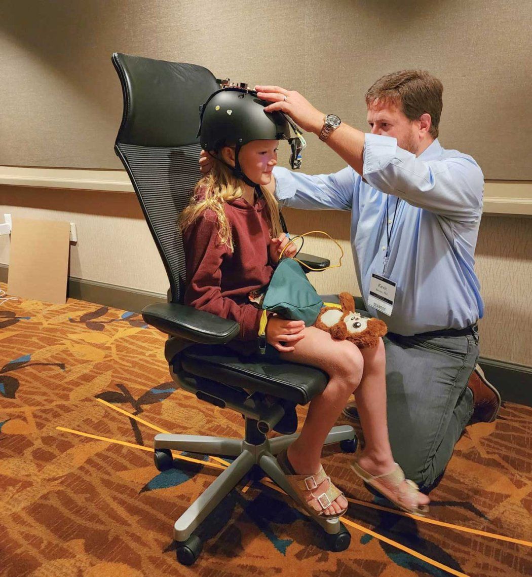 Kevin Bender (right) kneels as he adjusts a helmet on a young girl's head. She is sitting on an office chair, and the helmet will measure her eye movement.