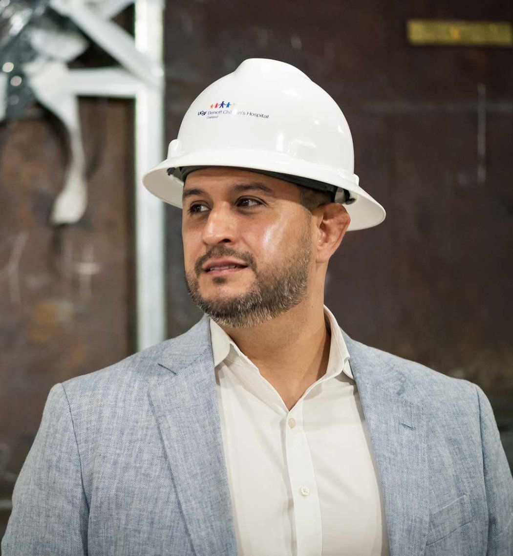 Construction project manager Michael Valero stands inside a construction zone and wears a white construction hat with the UCSF Benioff Children's Hospital Oakland logo on it.
