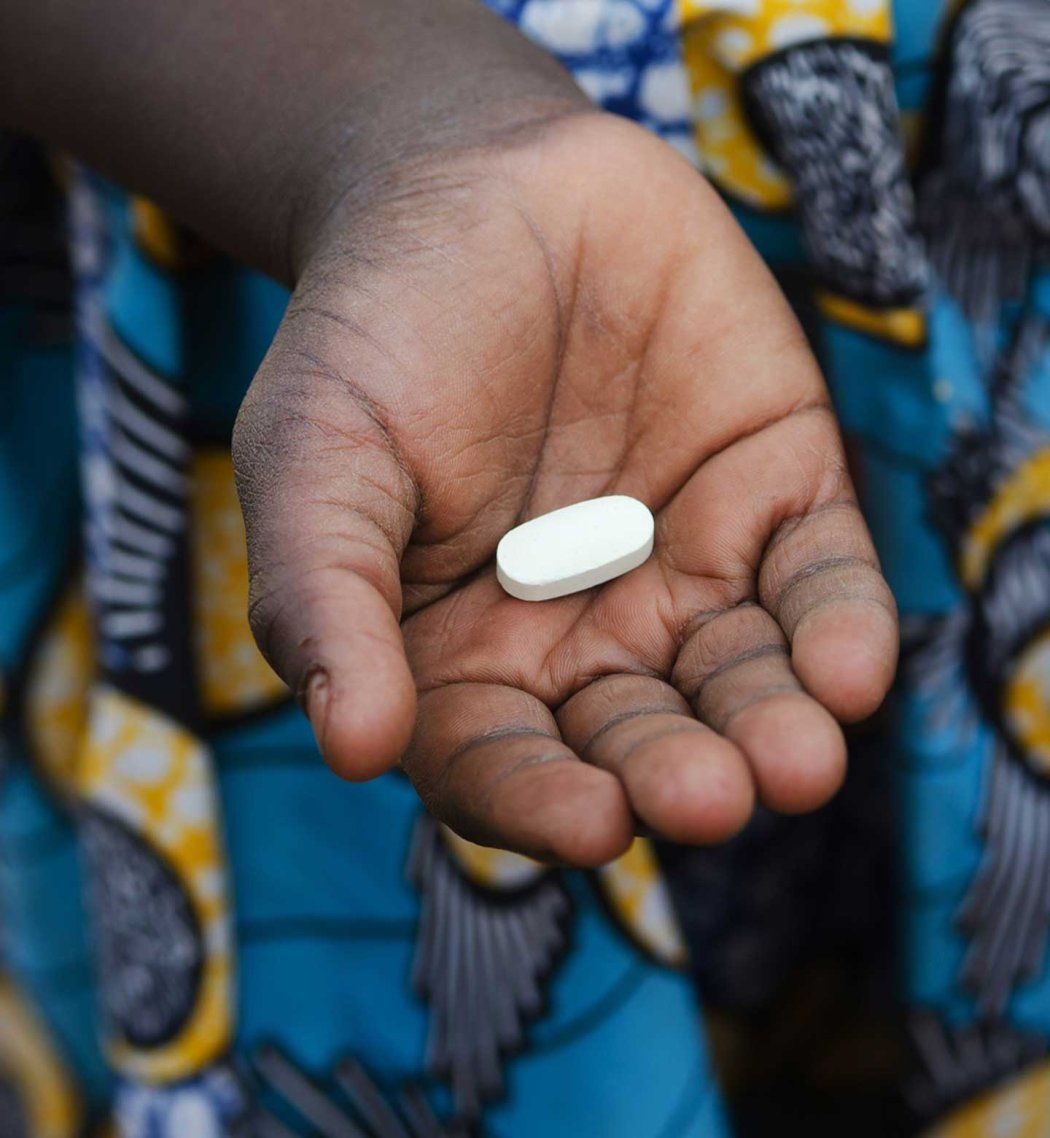 A close-up of a child's hand holding a single, oval-shaped white pill. The child is wearing a brightly patterned blue and yellow fabric.