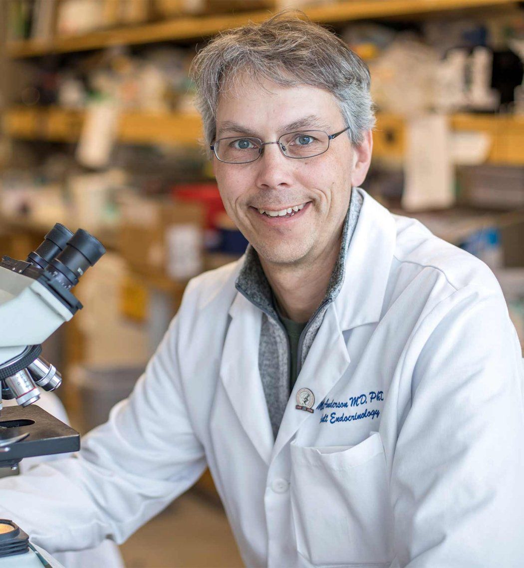Mark Anderson wears a white lab coat and sits in front of a microscope.