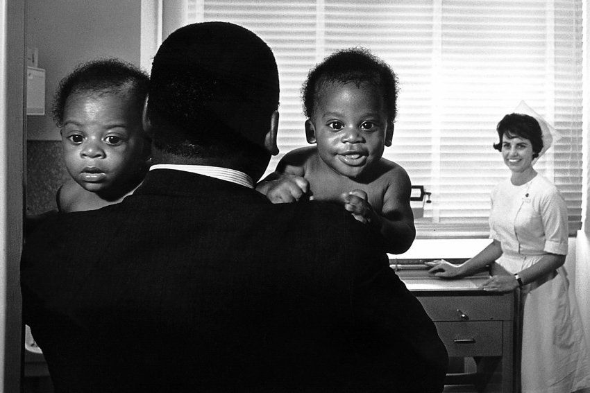 man carries his twin babies into an exam room where a nurse is waiting