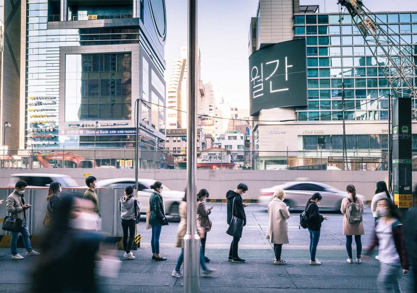On a busy street in a city in South Korea, people stand in line, several feet apart, with masks on, going about their daily lives.