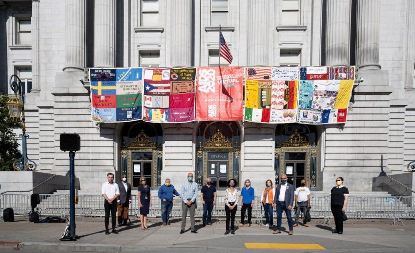 A colorful patchwork quilt hangs from city hall with various AIDS leaders and activists standing beneath it