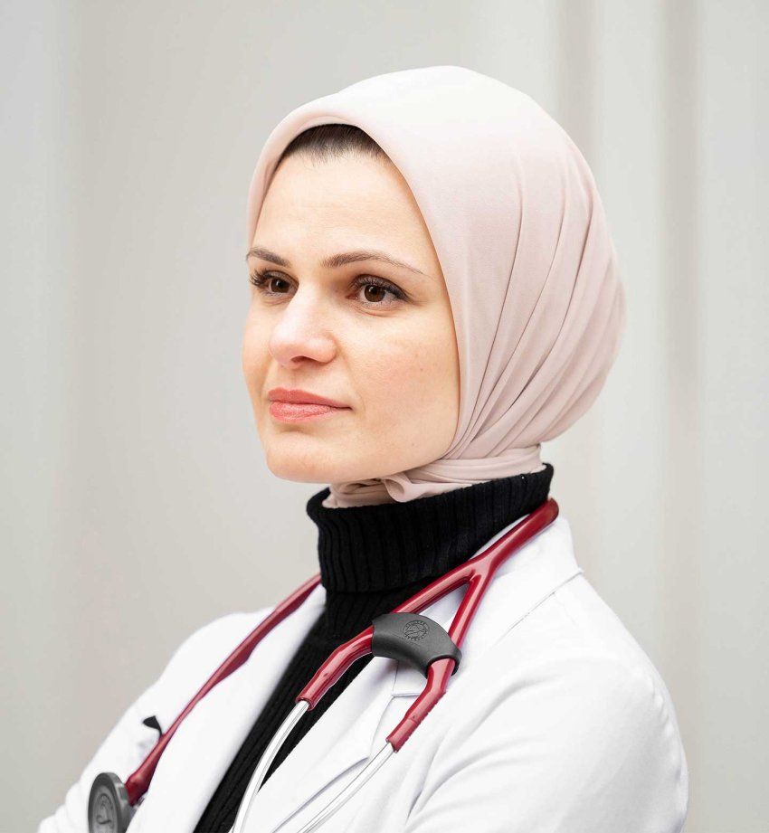 A portrait of Suzanne Barakat. She wears a white doctor's coat, a red stethoscope, and a pink hijab.