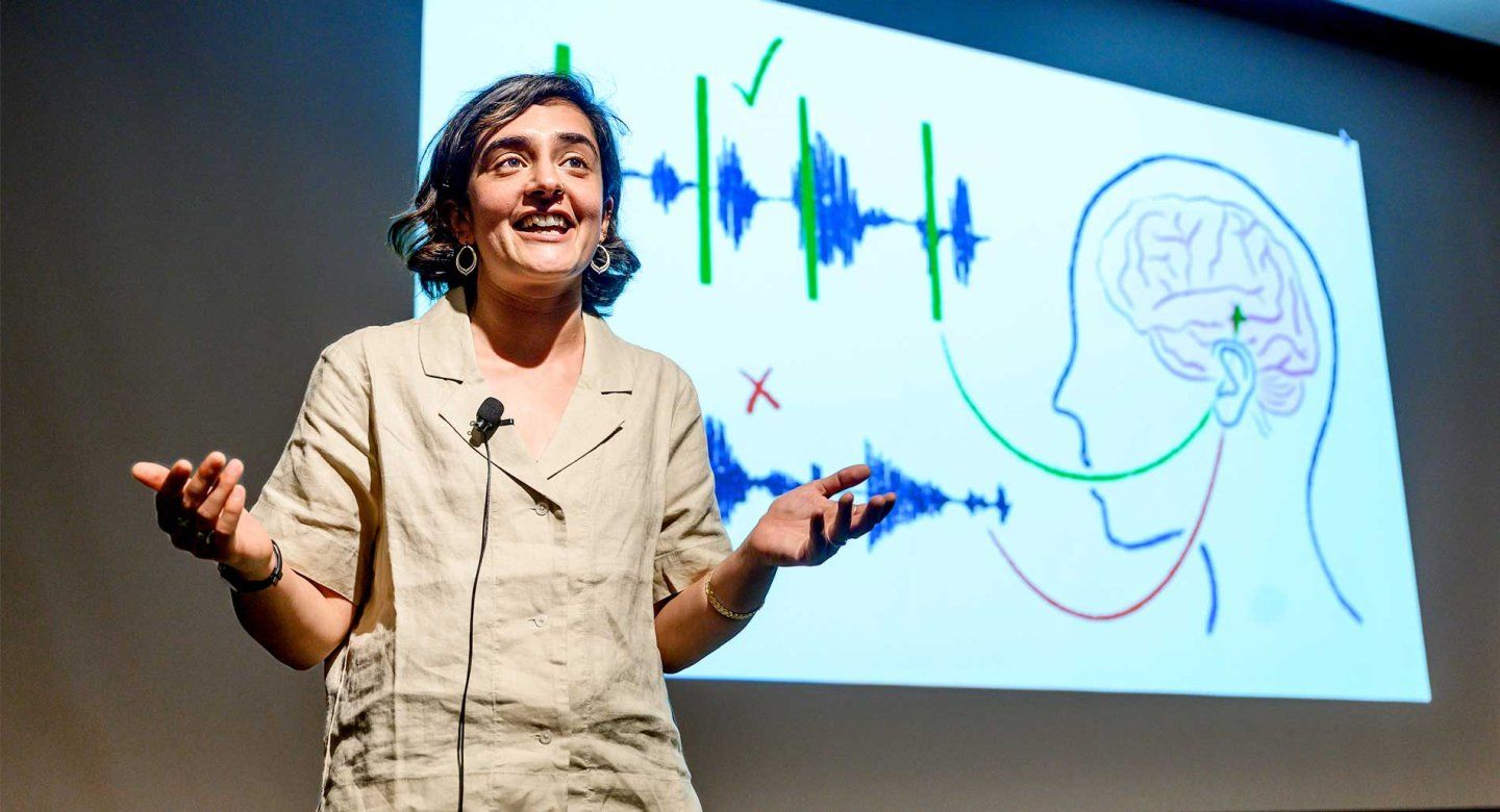 2024 Grad Slam winner Ilina Bhaya-Grossman presents her research onstage. In the background is a presentation slide showing an illusration of measurements of brainwaves.